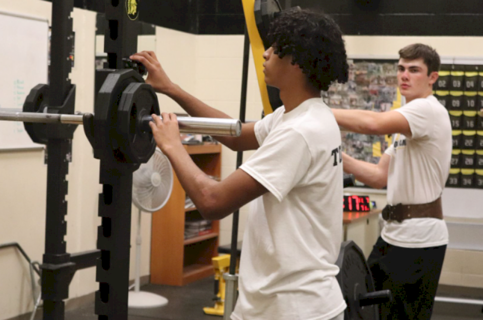 ADDING UP - Freshman Ethan McNeil,adds extra weight to the bar. Jake Wales watched as a reference to how much he should lift too. This day, the weights class was maxing out their back squat.