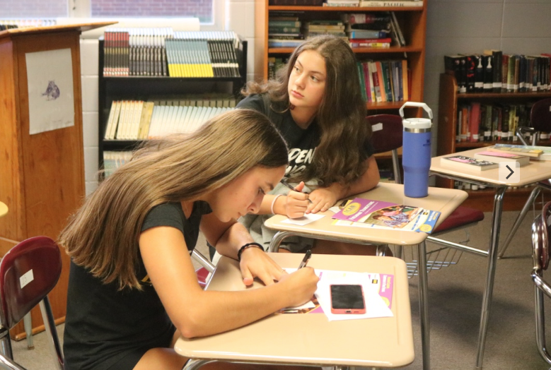 Freshmen Claire Comes and Saige Coffman write on slips of paper. Signing these slips of paper were a common practice for those entering teacher Ms. Berryhill's classes for the first time, and were a method to learn more about her students.