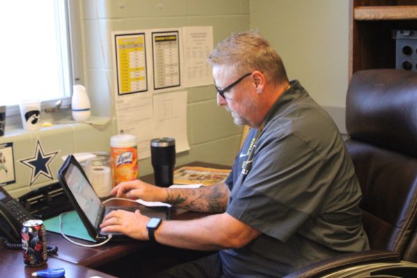 Derek Handel works diligently at his desk. Students surround him, receiving help with their homework. He works with them to get a better understanding of their work. 